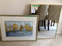 Framed watercolor picture by Susan Davis with natural rock formations rising from the water, adjacent to gold framed mirror, measurements visible on green tape.
