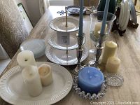 View of white tiered trays, various pillar candles including large blue candle, silver candle holders with blue taper candles, and glass hurricane candle holder on table.