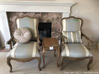 Overall view of two striped accent chairs with pillows flanking a carved wood side table against a fireplace