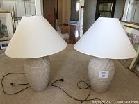 Two cream textured ceramic table lamps with white conical shades side by side on a carpeted floor. Black power cords visible.
