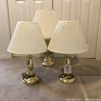 Three gold-colored base table lamps with white pleated shades arranged on carpeted floor.