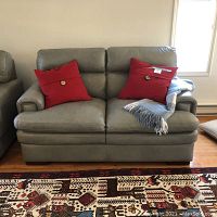 Front-facing view of the grey leather two-seat couch with two red pillows and grey throw on a hardwood floor.