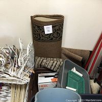 Photo showing three rolled entry carpets behind multiple clothes hangers, gray bucket, and brown baking pan.