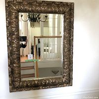 Large rectangular mirror with ornate gold frame showing full front view. Background includes stairwell and chandelier reflection.
