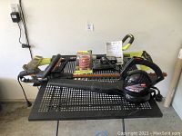 Front view of Ryobi hedge trimmer, Toro blower, battery charger, replacement line spool, and black metal table.