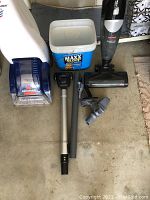 Photo includes Bissell Quick Steamer Plus steam cleaner, Bissell Magic Vac handheld vacuum, multiple vacuum attachments, and a Maxx Scoop cat litter box on concrete floor.