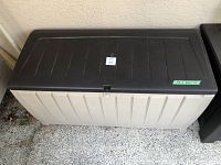 Large outdoor plastic storage bin with black lid and beige vertical panel body shown from the front.