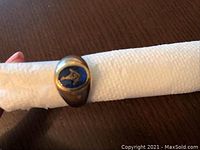 Front view of ring showing oval blue central stone with Masonic square and compass emblem set in yellow gold bezel, placed on white cloth over wooden surface.