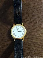 Full front view of watch showing gold case, white dial, Roman numerals and black leather strap