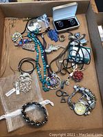 Overview of assorted costume jewelry items in a box including necklaces, bracelets, pendants, a pair of cufflinks, and brooches.