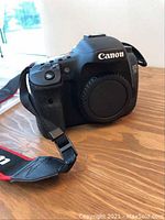 Front view, Canon EOS 7D body with body cap and attached Canon neck strap on wooden surface.