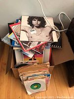 Cardboard box filled with a stack of LP album cover sleeves and a pile of 45 rpm records in paper sleeves on hardwood floor.