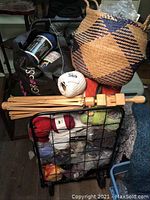 Photo showing a metal wheeled cart loaded with multiple balls of yarn, wooden knitting needles, a woven basket handbag, and a black tote bag containing more yarn accessories.