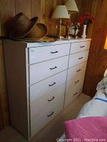 Front view of white 10 drawer dresser with black handles, top surface holding hats and lamps. Wood panel wall behind.