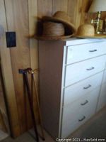 Two wooden walking canes with carved and curved handles leaning against wall beside dresser with straw hats on top