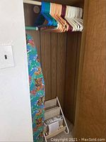 Closet area showing assortment of multi-colored plastic clothes hangers on a rod above an ironing board and a wooden magazine rack on the floor.