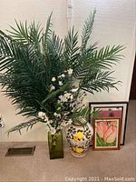Faux plant with white flowers in green container beside pottery urn with lemon design and two framed wall art paintings leaning against the wall.