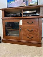 Front view of laminate TV cabinet showing glass door, drawers, and open shelves with electronic devices inside.