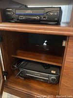 Panasonic VCR in black with multiple control buttons on front panel, stored in a wooden cabinet shelf with an Emerson VCR placed below it with visible VHS cassette slot and control dials.