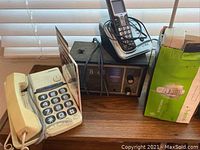 Lot of older small electronics including a vintage black analog clock radio, two beige corded telephones, a cordless phone on its charging base, and the original packaging box for a GE 900 MHz cordless phone.