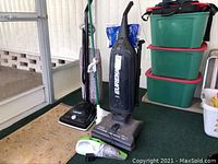 Photo showing three vacuum cleaners and one green and white squeeze handle mop all standing on floor next to storage containers, establishing lot contents.