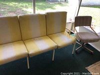 Full view of weathered outdoor couch with vinyl straps and yellow/white cushions, alongside a brown striped cloth swivel chair on green carpet indoors.