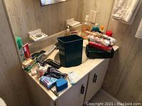 Photo of bathroom counter with assorted personal care products including razors, toothpaste, bandage box, lotion bottles and a green trash bin.