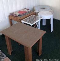 Photo showing five tables including a wooden table with glass top covered partially with fabric, small white metal mesh outdoor table, wooden side table, and two white plastic outdoor tables.