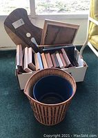 Photo of multiple wooden picture frames stacked inside a cardboard box with the largest frame leaning on the box side and a brown and black woven basket with a blue pot inside in the foreground.