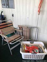 Two outside lawn chairs stacked and leaning against a wall, foldable wooden frame, laundry basket with miscellaneous items, and part of the lawn umbrella pole visible.