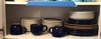Three blue cups next to multiple stacked white and blue plates and bowls on a shelf.