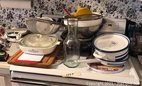 Kitchenware items on stovetop including Corning Ware casserole dish, enamelware pots, stainless steel strainers, glass carafe, and mixing bowls.