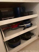 Three shelves inside a cabinet with various enamelware and metal pots, pans, and bakeware, including rectangular roaster, frying pans and small saucepans.