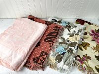 Four vintage tablecloths and linens folded and stacked side by side on a white surface against a white paneled background. One is pink damask, the others are vintage printed with floral patterns and fringed edges.