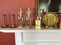 Photo showing the four pairs of brass candlesticks, three glass hurricane shades, and round brass serving dish arranged on a white mantel against a red wall.