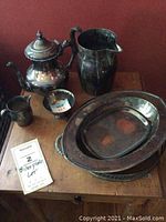 Full view of six silverplate pieces arranged on a wooden table showing pitcher, teapot, covered serving dish, tray, creamer, and small cup.