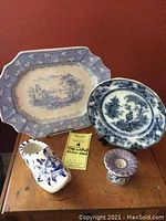 Four blue and white pottery items displayed on a wooden side table near a wall: large rectangular transferware serving plate, round Asian plate, Delft shoe ashtray, and Portuguese candlestick.
