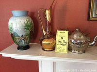 Front view of three items on a shelf against a reddish wall: painted glass vase, tall red Bohemian glass pitcher, and silver pottery teapot with lid on top