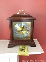 Front view of Howard Miller wooden mantel clock with brass and silver-tone dial showing Roman numerals, against a red wall background.