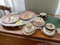 Wide shot of seven pieces of antique and vintage china arranged on table, showing variety of plates and cups with floral designs.