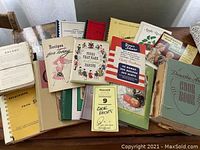 Wide view of the assortment showing multiple cookbooks stacked and spread across a table, including 'Recipes for Today,' 'Foods That Made New England Famous,' and 'The Domestic Arts Cookbook.' Also showing the Swans Down cake pan box.