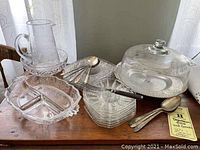 Full lot including glass pitcher, divided server, glass plates, cake dome on aluminum Lazy Susan, silver-plated spoons and fork on wooden surface.