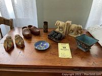 Photo showing an assortment of curios including carved wooden shoes, shell casing vase, stone elephant bookends, opera glasses, Chinese enamel pieces, and Belgium wooden shoe pin cushions on a wooden table.