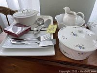 White ceramic casseroles and platters with floral designs, gravy boat with underplate, and rectangular platters arranged on a wooden surface, accompanied by a serving ladle and utensils.