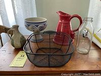 Front view of all items on wooden table under curtain light; two pottery pieces, wire basket, and milk bottle visible.