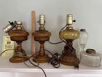 Three antique and vintage style lamp bases including two amber glass bases with intricate patterns and one greenish amber glass base. Electric sockets and cords are fitted. Extra glass parts such as a burner and a font are included.