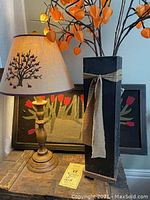 Full view of wooden lamp with embroidered lampshade, square black wooden floor vase with tied fabric bow holding orange light-up Chinese lantern branches, and appliqué tulip basket in background.