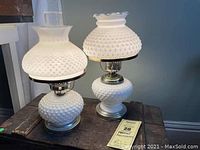 Two milk glass hobnail electric hurricane lamps side by side on wooden surface, one taller with ruffled top chimney glass, one shorter with simpler chimney.
