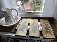 Three vintage botanical prints displayed on a wooden surface next to the pitcher and wash bowl