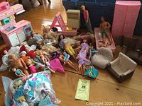 Photo of a large assortment of dolls including Barbie, Ken, Kelley, and Disney princesses, along with doll furniture pieces like a pink plastic kitchen and wardrobe, and seating furniture in brown and beige. Various dolls are in different states of dress. The floor around them is wooden.